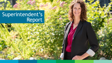 Woman with brown hair standing in front of green bush on a sunny day.