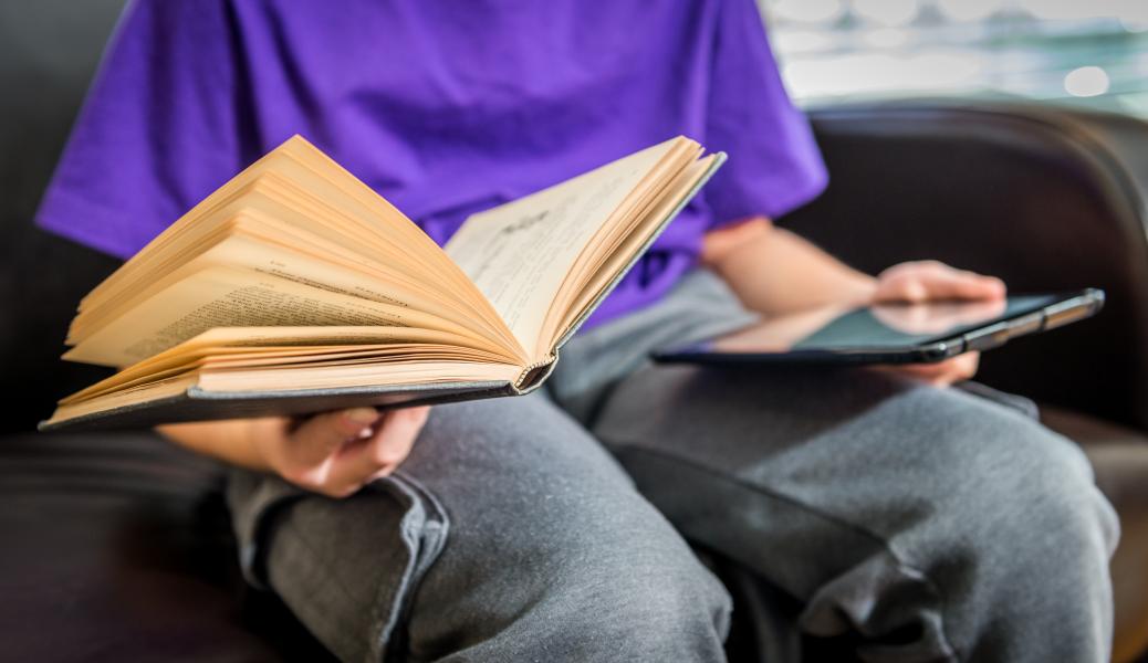 Child in purple shirt and grey jeans sitting holding an open book in one hand and tablet in the other.