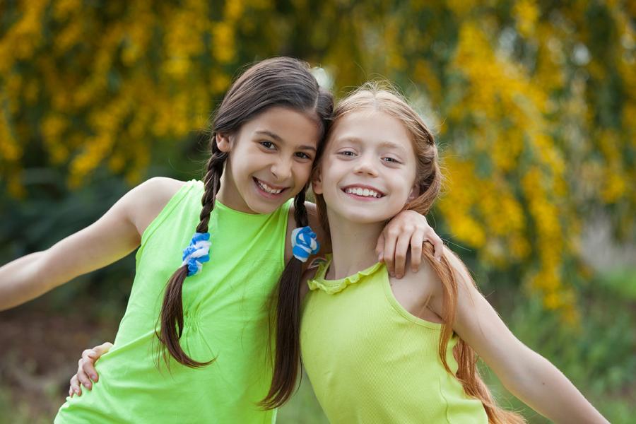 Two young girls wearing bright green and yellow sleeveless shirts and smiling broadly with arms around one anothers shoulders outdoors